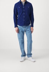 Blå kordflodsskjorta med två framfickor, kombinerad med ljusblå denimjeans och vita sneakers. Kläderna har en avslappnad passform och knappstängning.