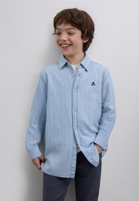 Hellblaues Hemd mit Knöpfen und vertikalen Streifen, langen Ärmeln und einem kleinen gestickten Totenkopf-Logo. Wird über einem weißen Hemd und dunklen Hosen getragen.