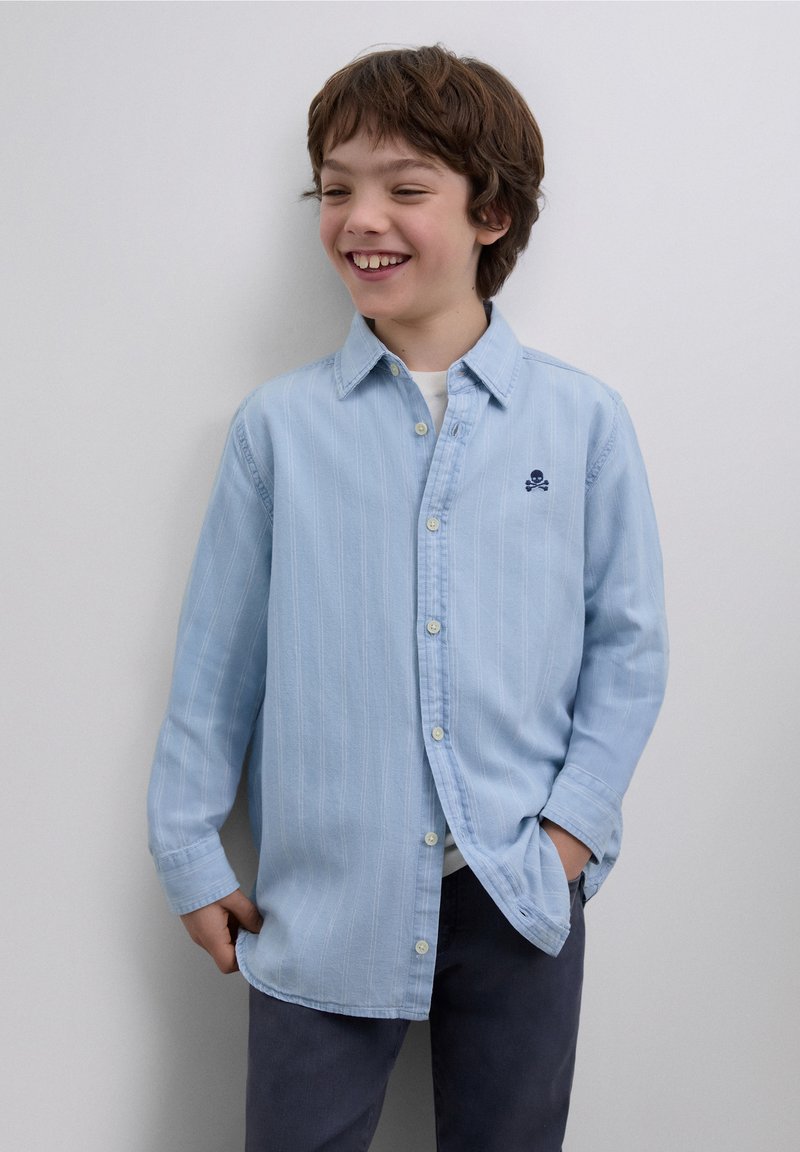 Hellblaues Hemd mit Knöpfen und vertikalen Streifen, langen Ärmeln und einem kleinen gestickten Totenkopf-Logo. Wird über einem weißen Hemd und dunklen Hosen getragen.