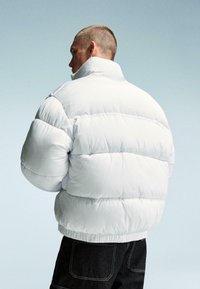 Vit puffjacka med hög krage, quiltat mönster och elastiska manschetter. Baksidan har minimal logotyp och är ihop med mörk denimbyxor.