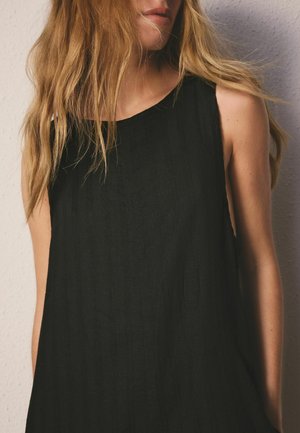 Blonde Frau in einem ärmellosen schwarzen, strukturierten Kleid, stehend vor einer hellen Wand, Haare locker über die Schultern gelegt.