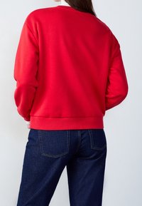 Mujer vistiendo un sudadera roja brillante de manga larga y jeans oscuros, de espaldas con las manos en los bolsillos contra un fondo claro y liso.