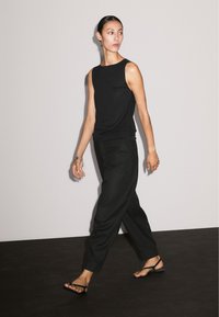 Een vrouw in een zwarte mouwloze top en losse zwarte broek loopt vol zelfvertrouwen, blootsvoets, tegen een effen lichte achtergrond, en toont een moderne stijl.
