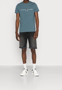 Tyrkysové bavlněné tričko s bílým logem, v kombinaci s tmavými džínovými šortkami se ohrnutými manžetami a bílými teniskami s černými akcenty.