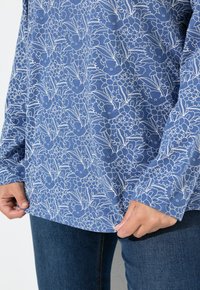 Camicia a maniche lunghe blu con motivo floreale bianco, indossata con jeans blu scuro, mani che tengono delicatamente l'orlo della camicia.