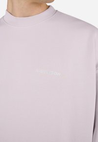 Light purple sweatshirt with a ribbed collar and a subtly embroidered "PEGADOR" logo on the chest. Soft texture and relaxed fit.