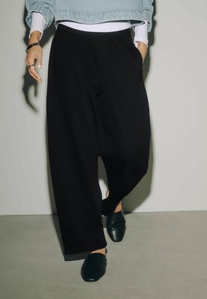 Kvinde går iført løstsiddende sorte bukser, sorte slip-on sko, cropped lys denimjakke og hvid skjorte.