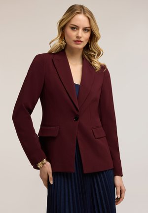 Kvinde med blondt hår iført en bordeaux enkeltknappet blazer over et navy plisseret skørt, accessoriseret med guldringe og armbånd.