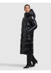 Lång svart vadderad jacka med glansig yta, huva och fickor med dragkedja, med vertikal sömnad och sidopartier. Bärs med svarta knähöga stövlar.