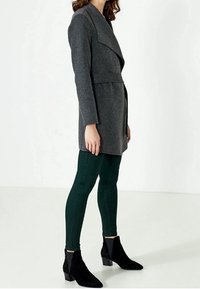 Osoba oblečená v přiléhavém tmavě šedém kabátu s páskem, tmavě zelených skinny kalhotách a černých kotníkových botách, stojící na jednobarevném bílém pozadí.