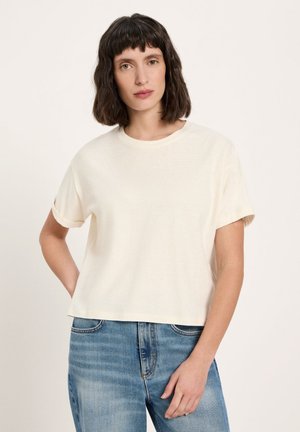 Woman with short dark hair wearing a plain off-white short-sleeve t-shirt and blue jeans, standing against a light background.