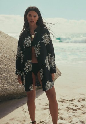Woman wearing black floral kimono over black bikini standing on sandy beach near large rock with ocean waves in background.