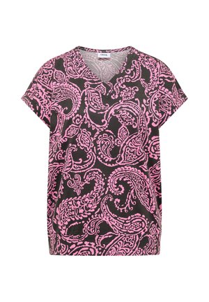 Femme souriante marchant, portant un chemisier rose à motifs, un pantalon beige, des chaussures marron et un sac cabas noir texturé dans une pièce neutre.