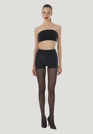 Schwarzes Bandeau-Top kombiniert mit hochtaillierten schwarzen Shorts und transparenten schwarzen Netzstrümpfen. Das Modell trägt schwarze Stöckelschuhe. Einfarbiger Hintergrund.