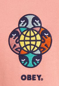 Vyšité logo na měkké růžové tkanině, které zobrazuje kruhový design se čtyřmi obličeji v zelené, modré, oranžové a růžové barvě, kolem žluté planety.