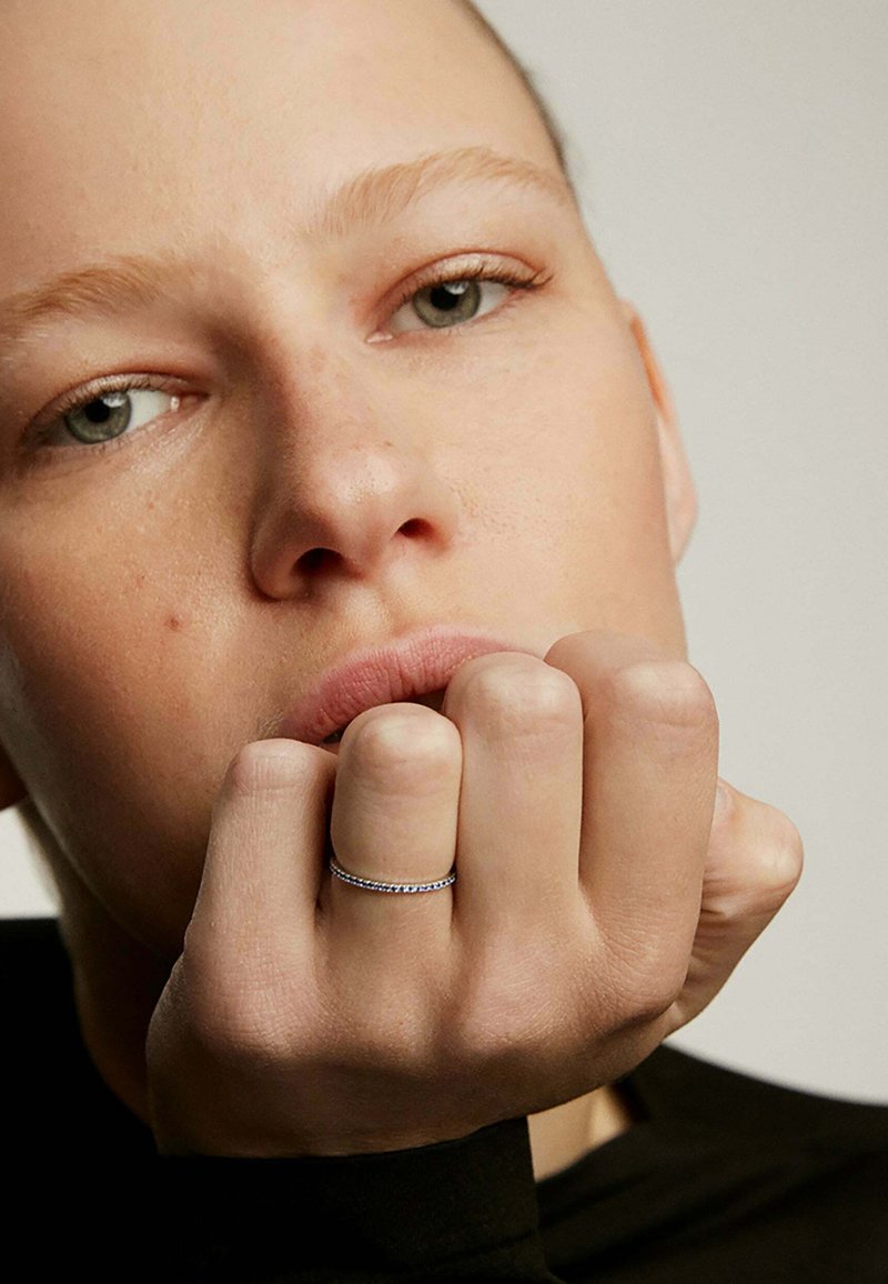 Anillo de plata con pequeñas piedras preciosas azules, llevado en el dedo anular, destacando la textura de la piel y la apariencia natural de las uñas contra un fondo oscuro.