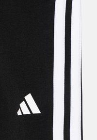 Pantalones atléticos negros con tres rayas verticales blancas y un logo blanco de Adidas en la parte inferior izquierda. Textura de tejido de punto.