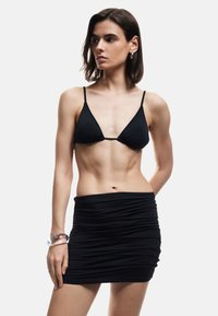 Czarny biustonosz bikini z trójkątnymi miseczkami i cienkimi ramiączkami, zestawiony z marszczoną czarną mini spódniczką. Przezroczysta bransoletka na nadgarstku. Gładka tekstura materiału.