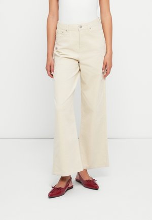 Mujer con pantalones anchos color marfil y zapatos planos slingback rojos de punta afilada, de pie frente a un fondo blanco.