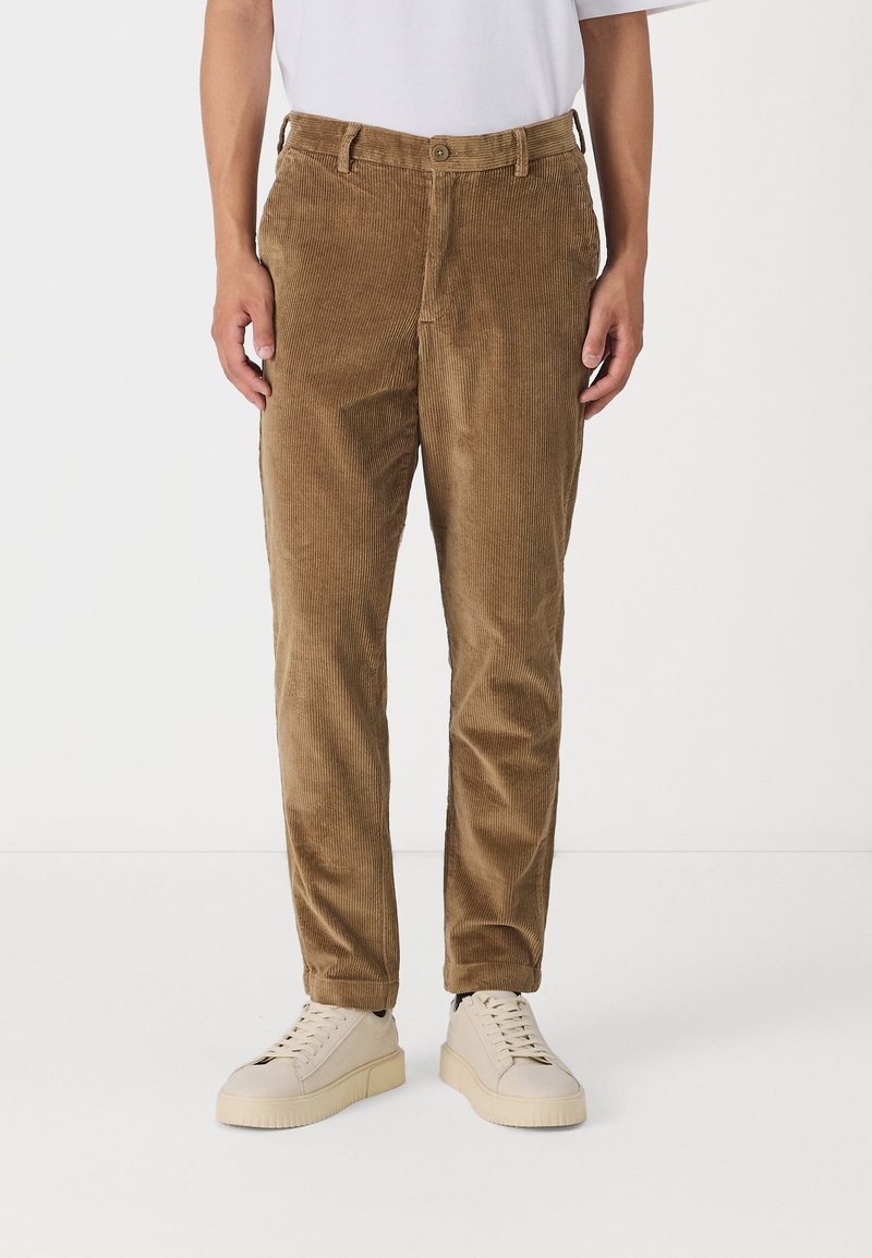Homme portant un pantalon en velours côtelé marron, des baskets blanches et une chemise blanche, debout devant un fond blanc uni.