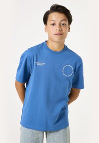 Camiseta de algodón azul con mangas cortas, escote redondo y detalles impresos en blanco en la parte delantera. Textura de tela natural y corte relajado.