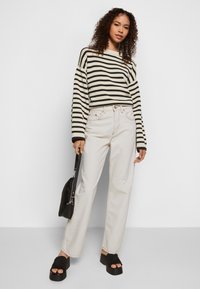 Sweater às riscas preto e creme, jeans de cintura alta em beige claro, sandálias plataforma pretas volumosas e uma mala de ombro preta.