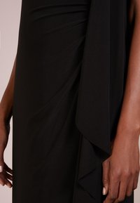 Svart klänning med draperat, flödande tyg med mjuk textur. Designen har samlade detaljer vid midjan och en lös ärmaccents.