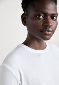 T-shirt blanche à manches courtes en tissu texturé avec un col rond, portée par une personne aux cheveux très courts et à la peau lisse.