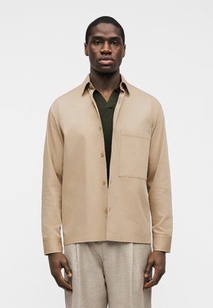 Man wearing beige button-up shirt over dark green V-neck sweater with light gray trousers against plain white background.