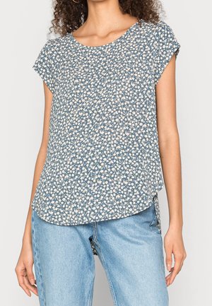 Kvinde iført en blå blomstret kortærmet top med rund halsudskæring og lyseblå denimjeans mod en ensfarvet baggrund.