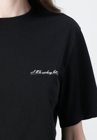 T-shirt in cotone nero con maniche corte e scollo tondo, caratterizzata da un dettaglio testuale ricamato bianco sul petto: "Lavorerò fino a tardi."