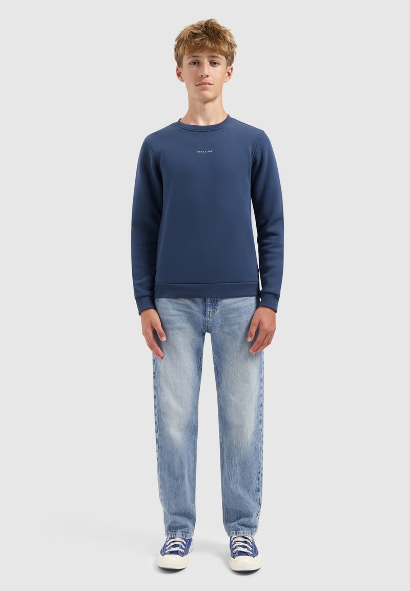 Tienerjongen staat stil in een marineblauwe sweatshirt, lichtblauwe jeans en blauw-witte sneakers tegen een plain achtergrond.