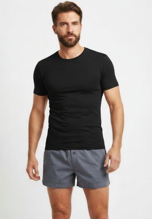 Bärtiger Mann steht, trägt ein eng anliegendes schwarzes T-Shirt mit kurzen Ärmeln und graue, geknöpfte Shorts vor einem schlichten weißen Hintergrund.