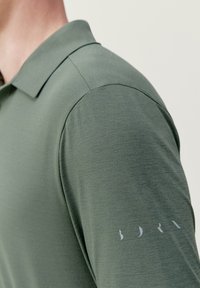 Polo en vert discret, présentant un tissu texturé et un col classique. Détail notable : logo gris clair sur la manche.