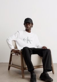 Pullover bianco con logo "Calvin Klein" grigio, pantaloni neri e scarpe da ginnastica nere. Modello seduto su una sedia in legno chiaro contro uno sfondo neutro.