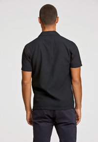 Maglietta nera a maniche corte realizzata in tessuto testurizzato, con colletto piatto e orlo dritto. La vista posteriore mostra cuciture minime e un design semplice.