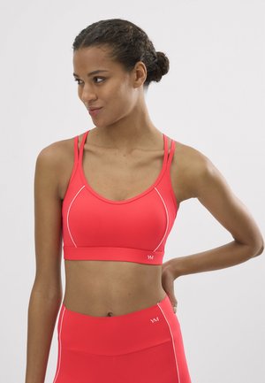 Mujer con el cabello oscuro recogido en un moño, vistiendo un sujetador deportivo rojo y leggings de entrenamiento a juego de cintura alta, mirando hacia su izquierda con la mano en la cadera.