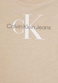 Beige puuvillainen t-paita, jossa suuret valkoiset "CK"-kirjaimet ja harmaat "Calvin Klein Jeans" -tekstit. Sileä pinta, klassinen pyöreä kaula-aukko.