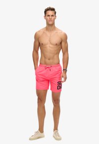 Neonroze zwemshorts met een trekkoord in de taille, met een groot zwart "SUPERDRY" logo op het linkerbeen. Lichtgewicht, gladde textuur.