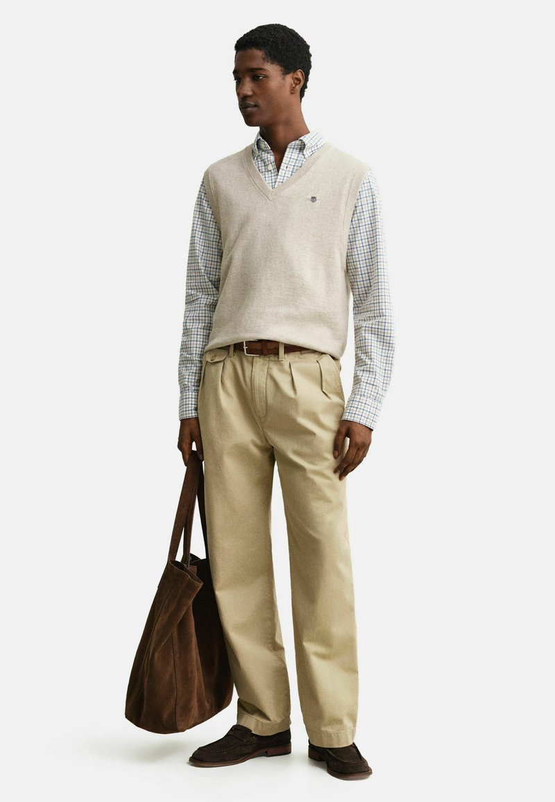 Jeune homme portant un pull sans manches beige par-dessus une chemise à carreaux avec un pantalon kaki, tenant un grand sac fourre-tout marron.