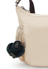Beżowa torba crossbody z tkaniny z kieszonką na zamek z przodu, z ciemnozielonym zawieszką w kształcie goryla wykonaną z miękkiego materiału, przymocowaną do breloczka.