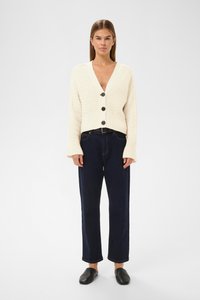 Kreemja ribakardigan sügava V-kaelusega, millel on kolm musta nööpi, koos tumedate teksapükste ja mustade sussidega.