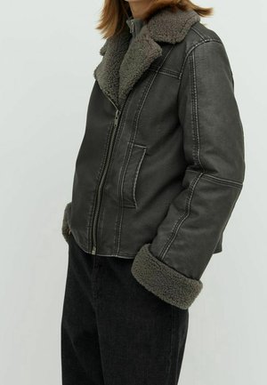 Person trägt eine dunkelgraue Lederjacke mit Pelzkragen und -bündchen, kombiniert mit schwarzen Hosen vor schlichter Hintergrund.