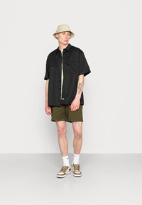 Zwarte kortekiel met knopen, twee borstzakken, groene T-shirt eronder, olijfgroene shorts en een beige bucket hat. Bruine sneakers aanwezig.