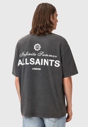 Mand med skulderlangt hår iført en falmet sort T-shirt med hvid tekst "Infinite Summer ALLSAINTS LONDON" på ryggen.