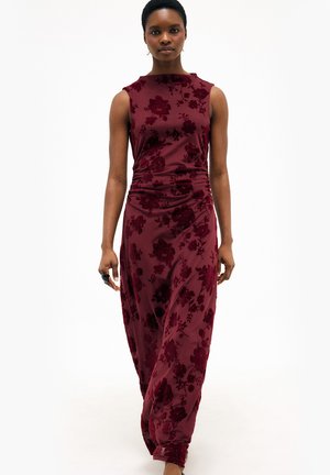 Woman walking forward wearing a sleeveless dark red floral maxi dress with ruched waist and holding a black ring on her right hand.