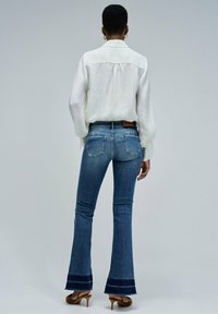 Jeans de mezclilla azul de campana con un dobladillo en azul marino oscuro, combinados con una blusa blanca de manga larga texturizada. Bolsillos traseros visibles y una etiqueta de cuero.