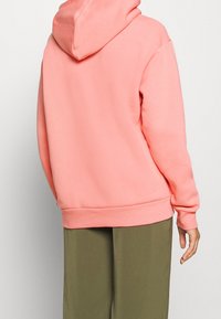 Mikina s kapucí v světle korálové barvě, vyrobená z měkké látky. Má žebrovaný lem a manžety, volný střih a minimální počet švů.
