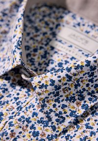 Camicia bianca con un motivo floreale blu, giallo e marrone, caratterizzata da un colletto classico e un tessuto testurizzato. Etichetta visibile all'interno del colletto.