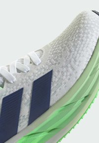 Running shoe featuring a textured white upper, blue branding stripes, and a green midsole with a rubber outsole for grip.
